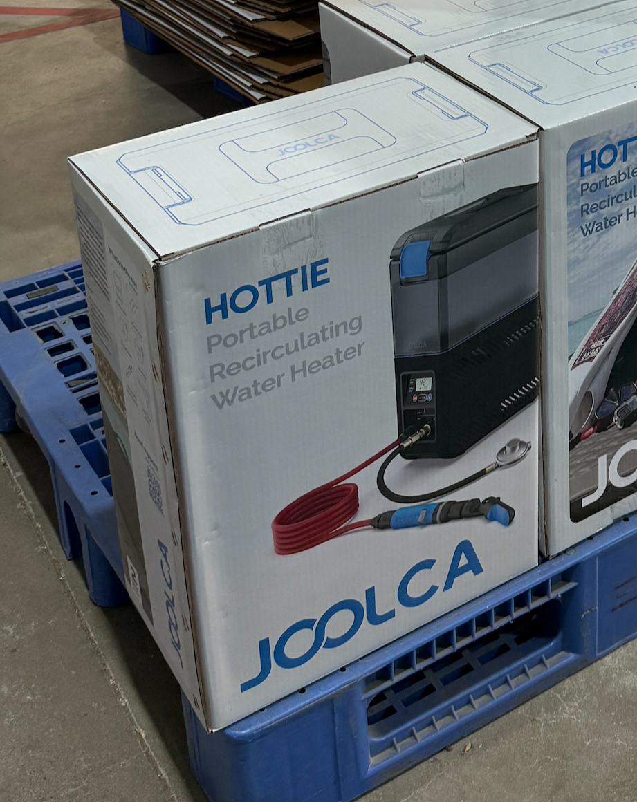 Stacked packaging boxes for a portable recirculating water heater system branded Joolca, designed for outdoor and off-grid use, shown on a blue pallet in a warehouse setting.