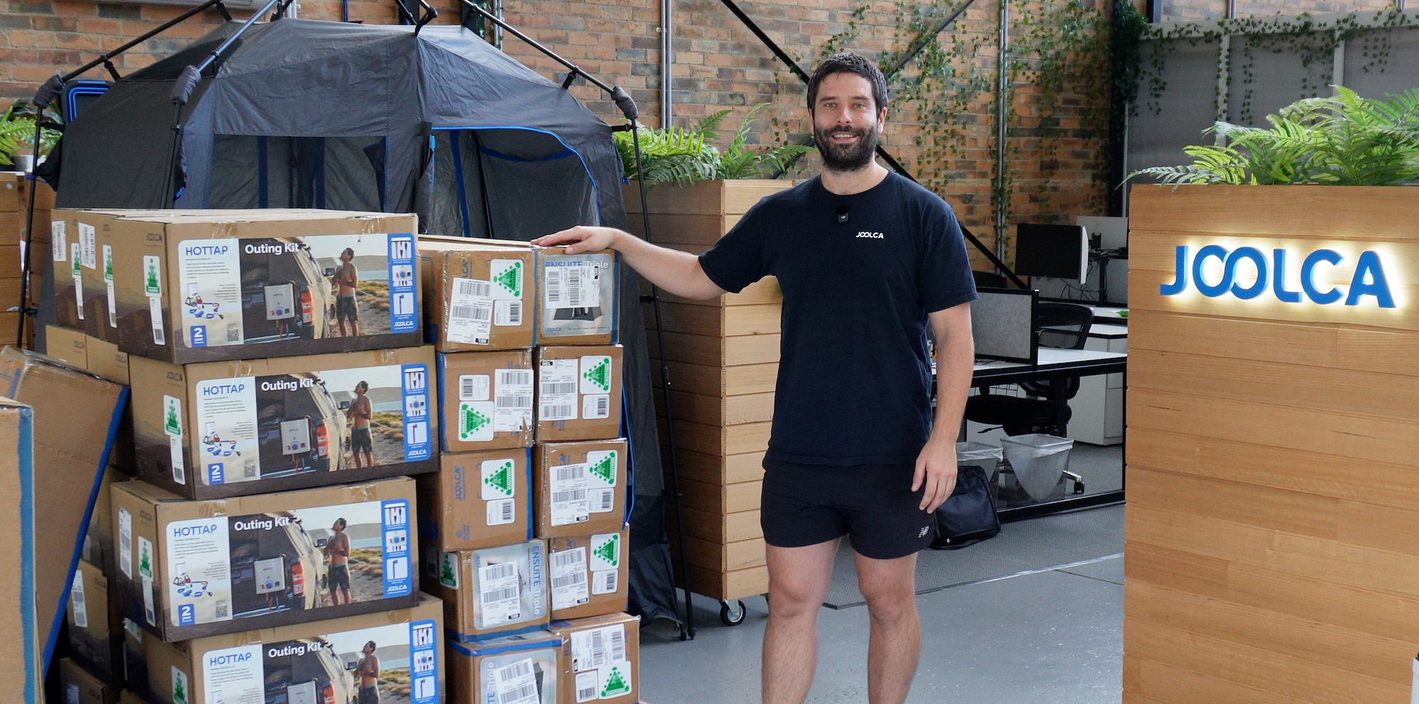 Man standing outdoors next to a stack of boxed camping hot water systems with a privacy tent and a wooden sign displaying the brand name Joolca in the background.