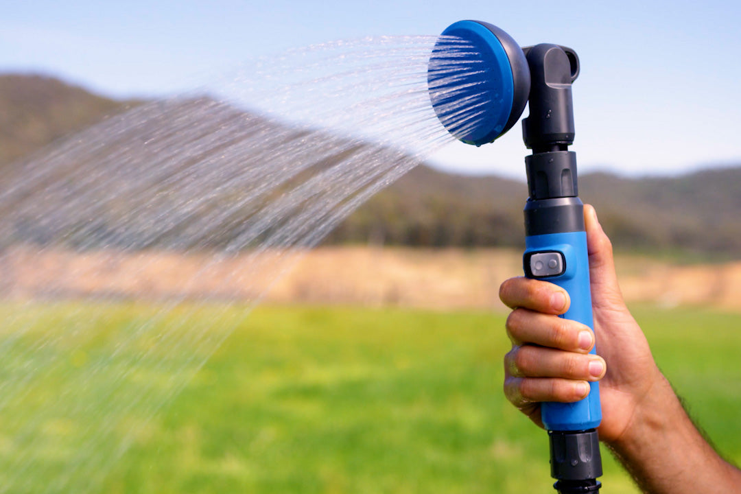 Hand holding the HOTTAP V2 shower head, spraying water over a green paddock