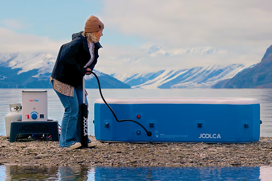 Person inflating a HOTTUB with a hand pump on a lakeshore, with a HOTTAP V2 nearby and snowy mountains behind