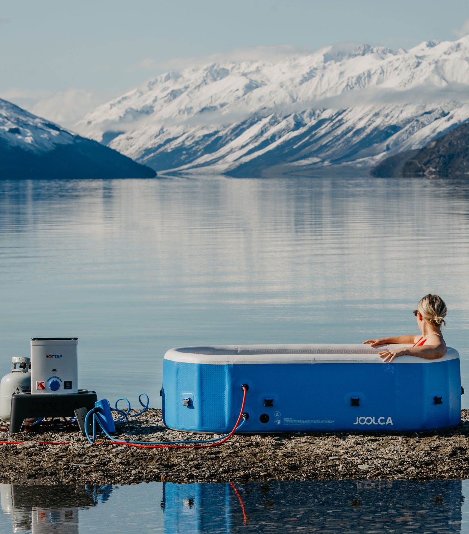 A person relaxing in a blue portable outdoor hot tub on a rocky lakeshore with snow-covered mountains in the background and a compact propane water heater setup nearby.