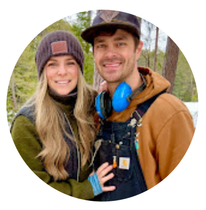 A smiling man and woman outdoors in a forested area, dressed in warm casual clothing including a beanie, hoodie, and overalls with blue ear protectors around the man's neck.
