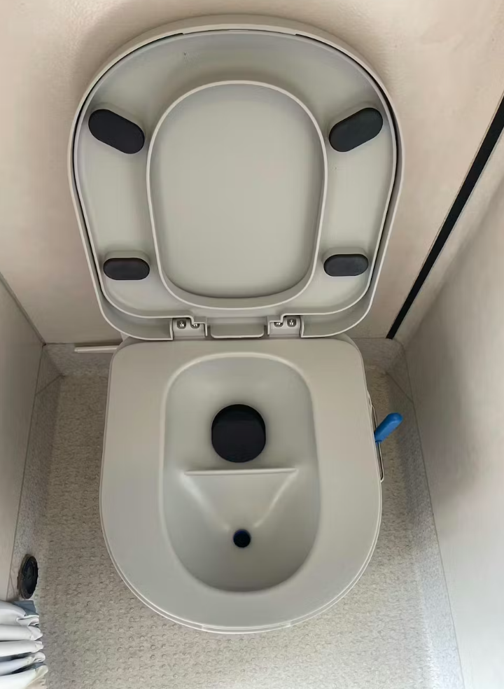 Top-down view of a portable composting toilet with an open lid inside a small enclosed space, featuring a simple, compact design and a blue lever handle on the side.