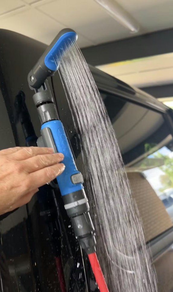 A portable outdoor shower head attached to a hose, spraying a steady stream of water, held by a hand against a dark vehicle surface in an outdoor setting.