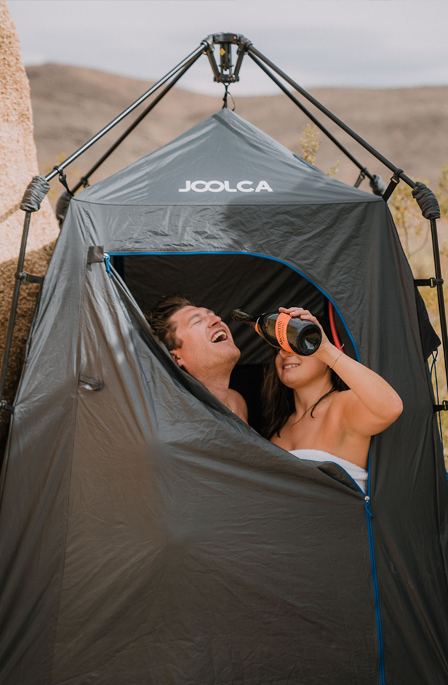 Two people inside a portable privacy tent branded Joolca, one laughing while the other holds a bottle, set in an outdoor, off-grid environment.