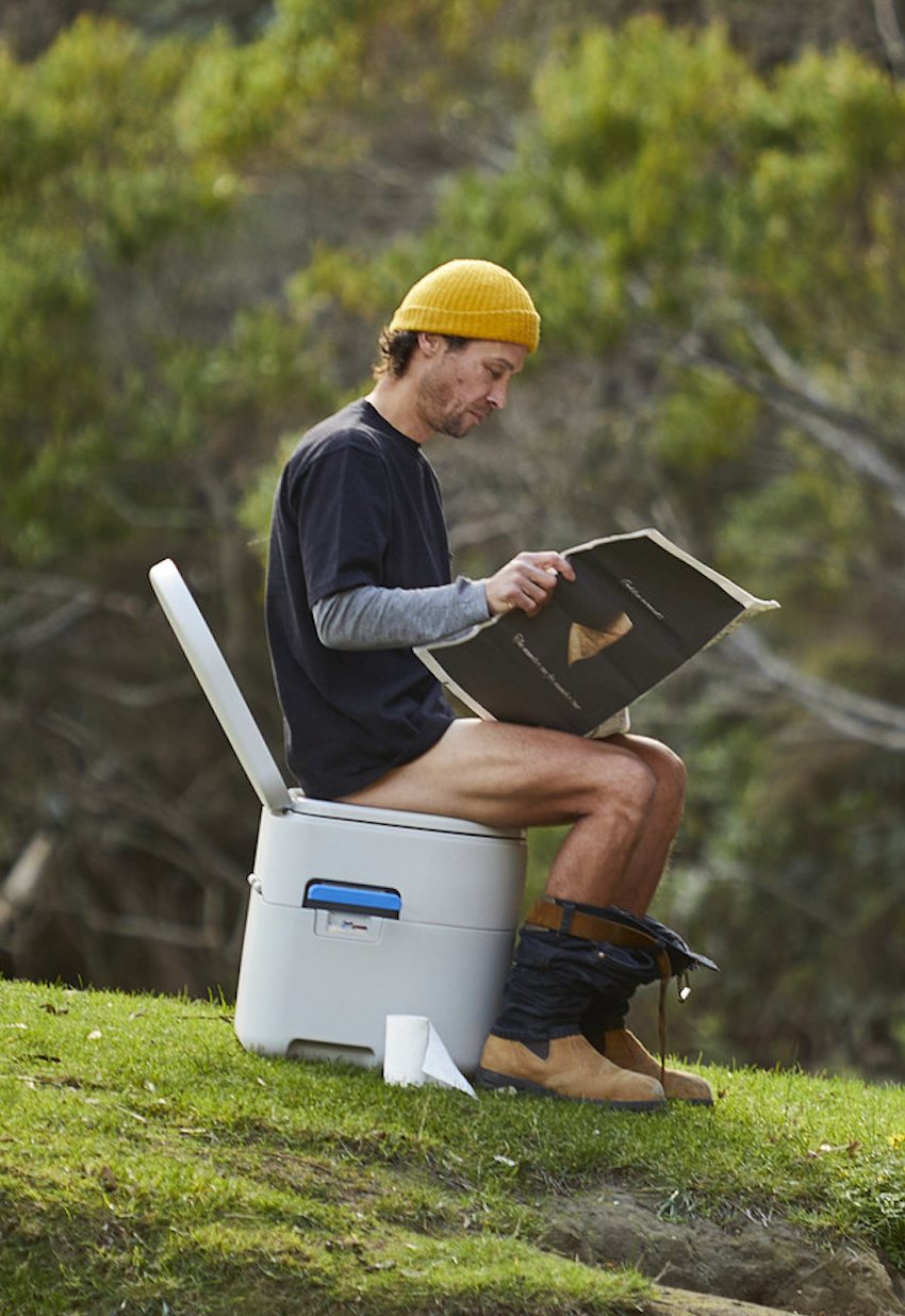 Person sitting on a portable toilet outdoors reading a book