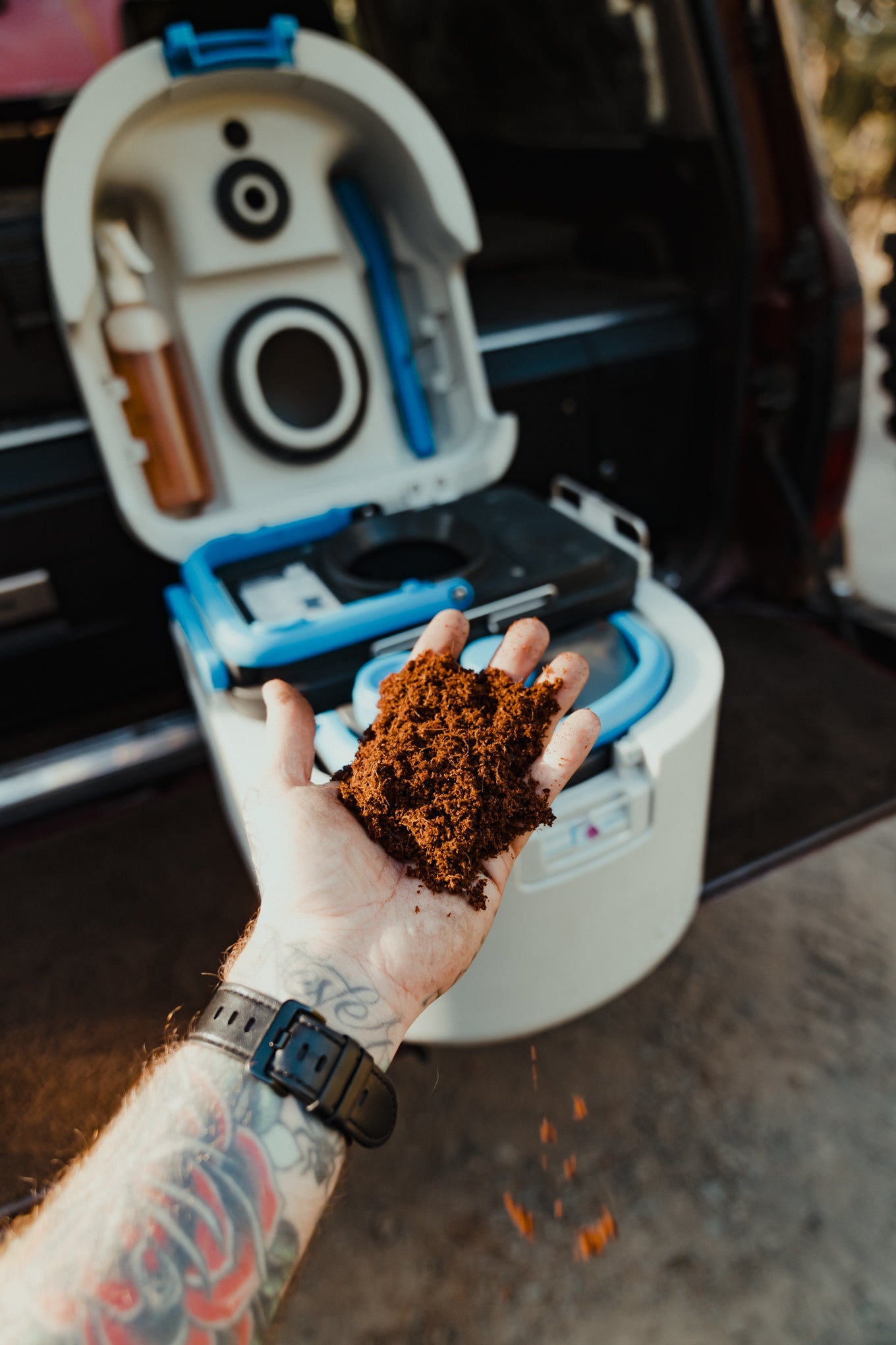 Hand holding coffee grounds in front of a portable toilet with tattooed arm and watch.