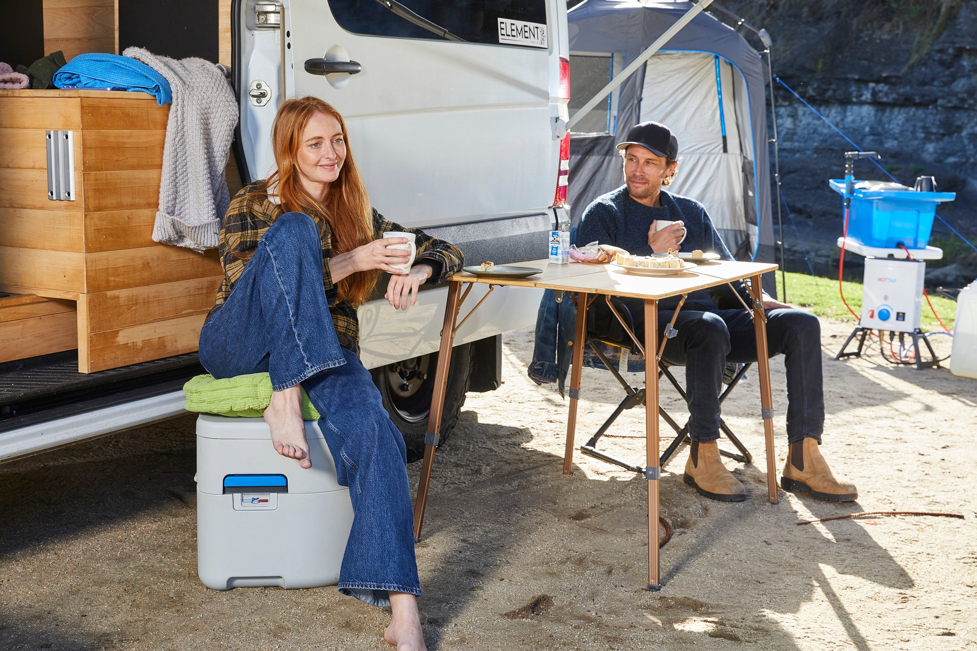 Two people sitting at a table outdoors near a camper van with a cooler box.